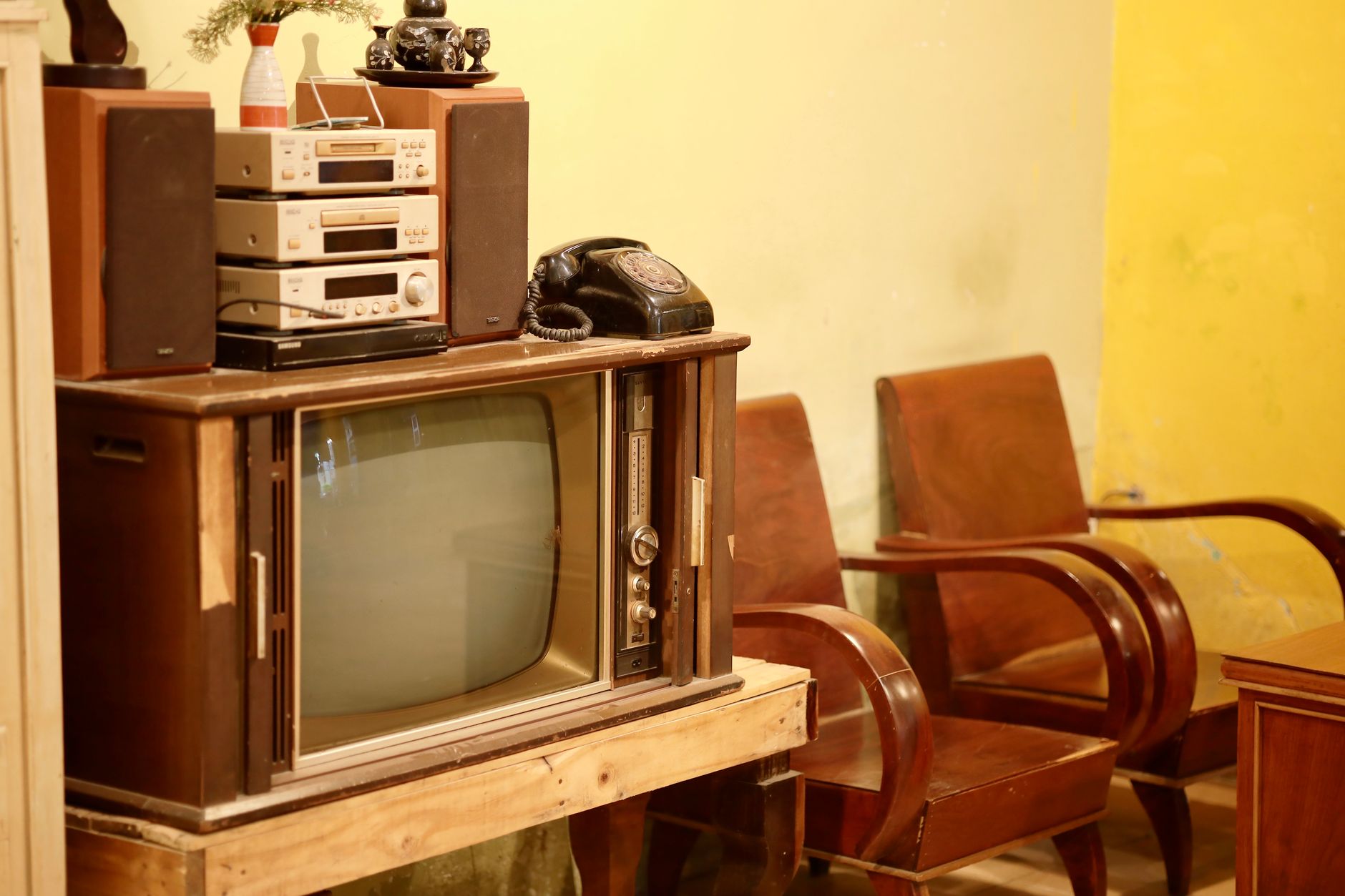 Ancien téléviseur dans un meuble en bois, avec chaîne hi-fi, enceintes et téléphone à cadran, près de deux fauteuils en bois.