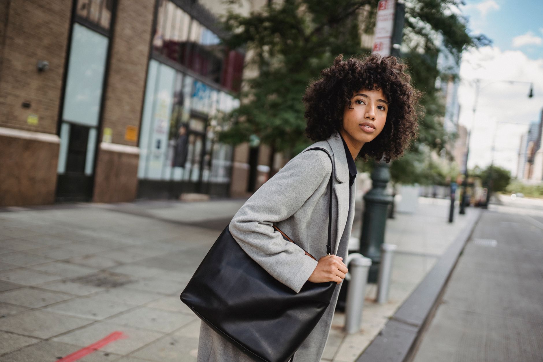 Femme aux cheveux bouclés en manteau gris, portant un grand sac noir en cuir sur l’épaule, sur un trottoir de ville.