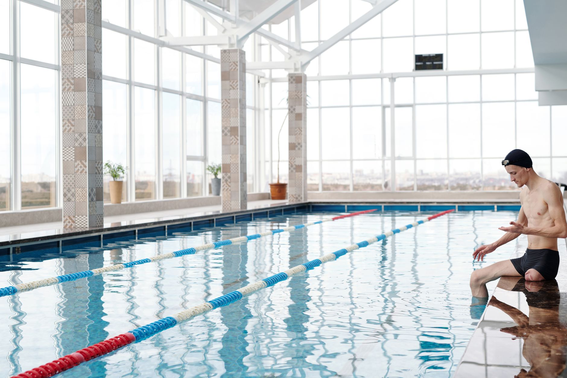 Homme en bonnet de bain assis au bord d’une grande piscine intérieure aux baies vitrées, avec des lignes d’eau rouges et bleues.