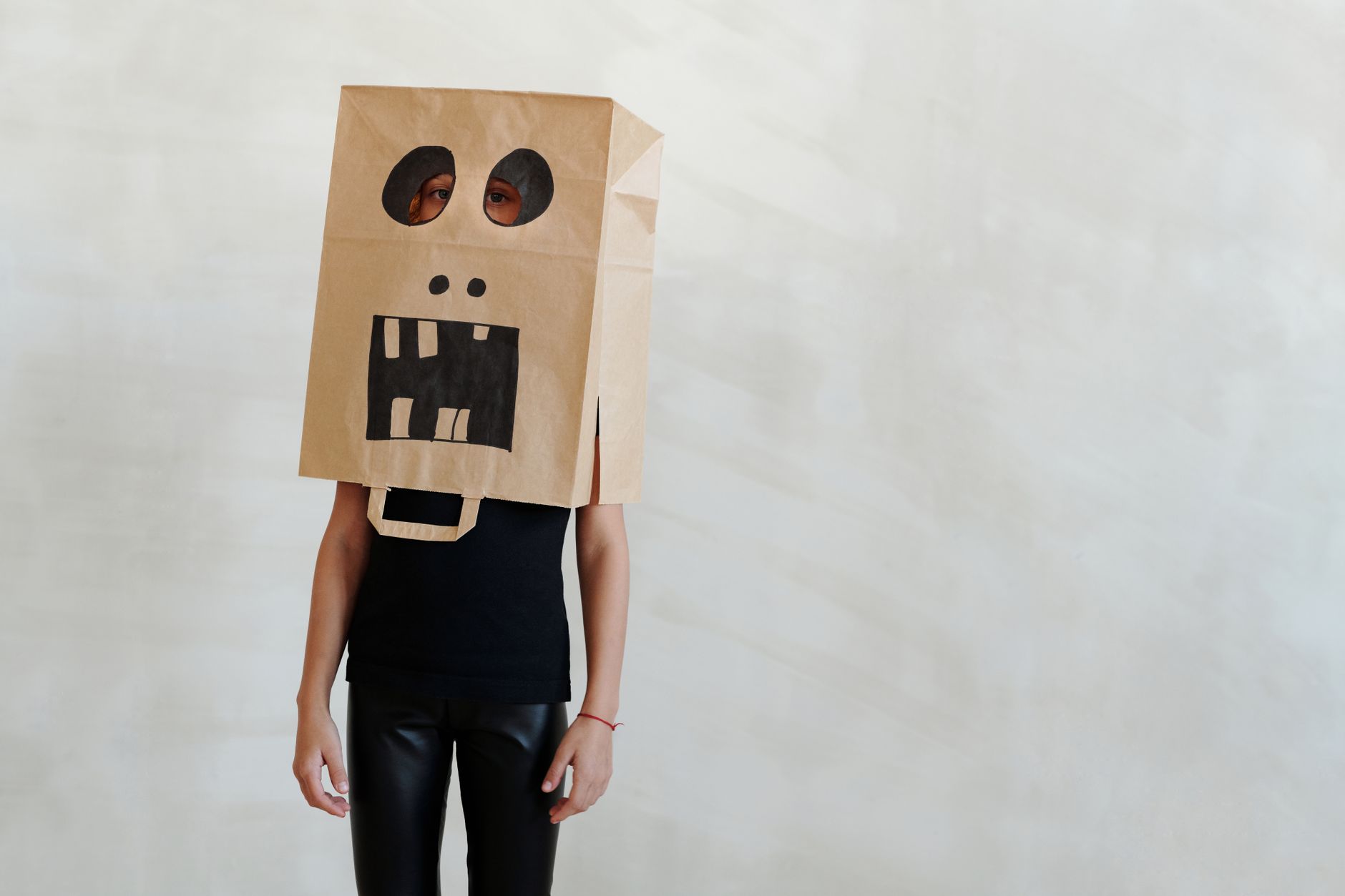 Personne debout avec un sac en papier kraft sur la tête, décoré d’un visage de monstre aux grandes dents, haut noir et pantalon noir brillant.