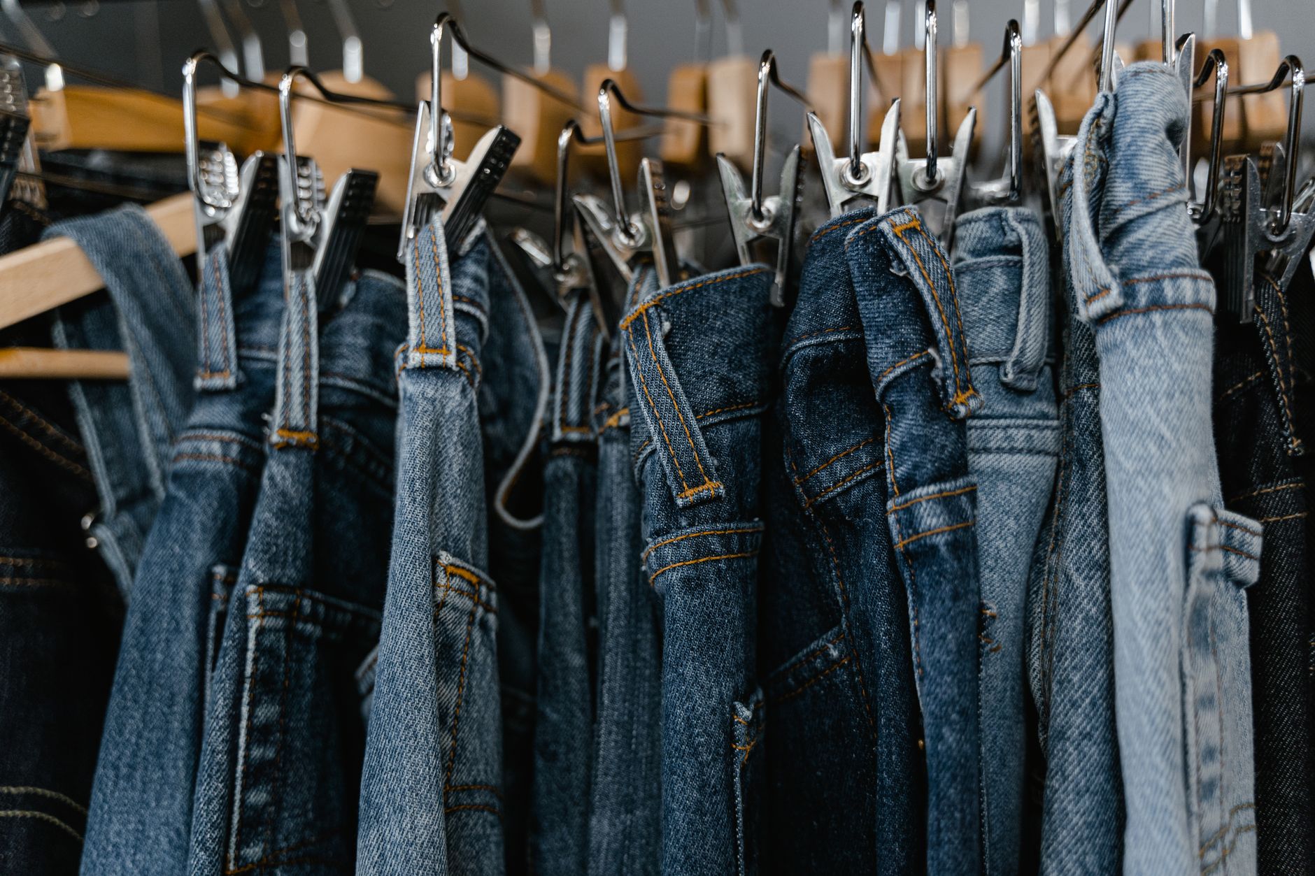 Rangée de jeans en denim bleu, du clair au foncé, suspendus à des cintres en bois avec pinces métalliques.