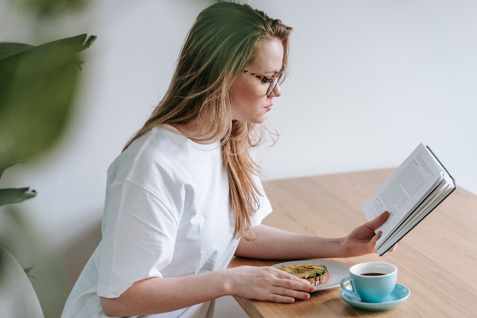 Une femme lisant un livre devant une tasse de café et une tartine sur une table en bois.
