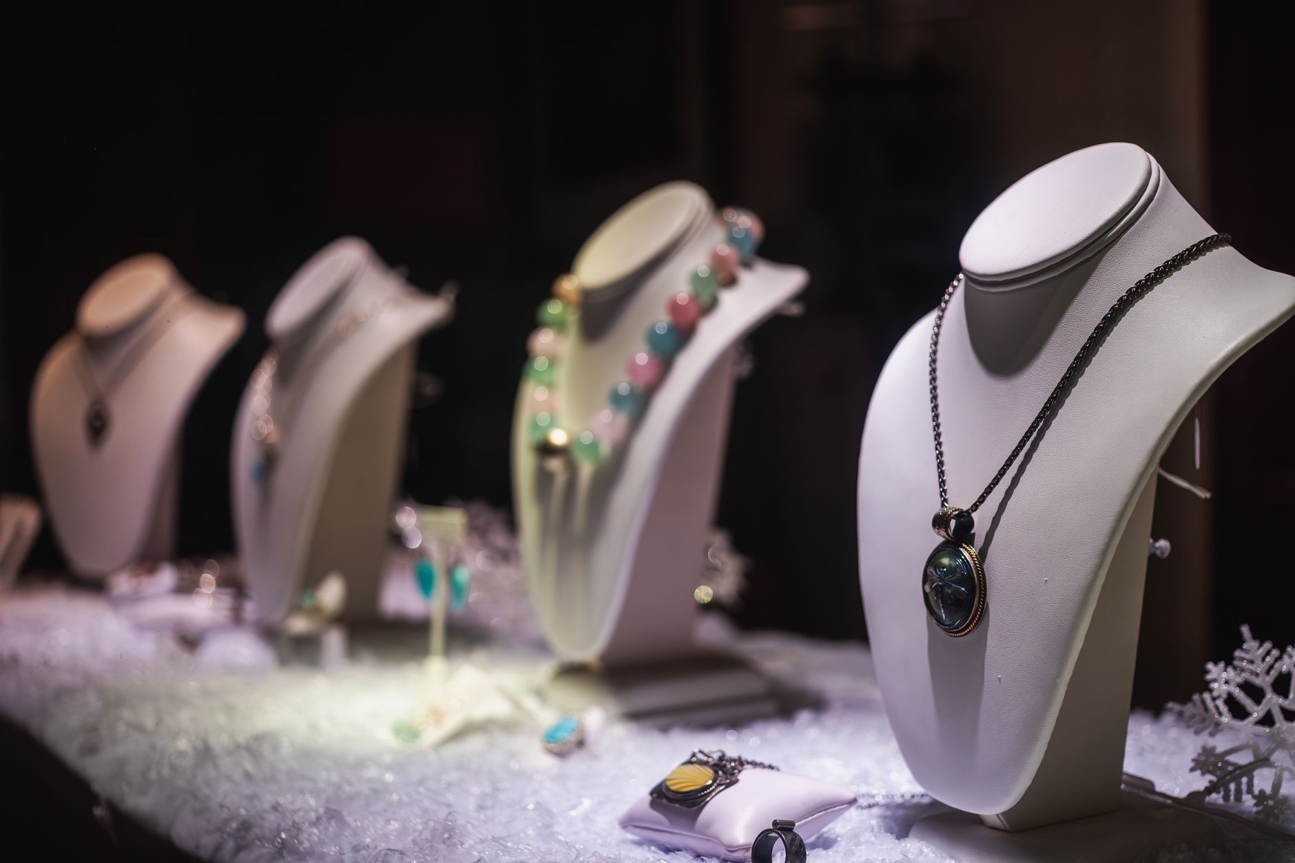 Vitrine de bijouterie avec des bustes blancs présentant des colliers, dont un pendentif bleu-vert et un collier de perles colorées, sur de la neige artificielle.
